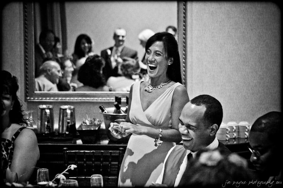 A black and white image of a woman laughing joyfully in a social gathering. Shes standing beside a seated man who is also smiling. Others are seated around a table, and a mirror reflects additional people in the room.