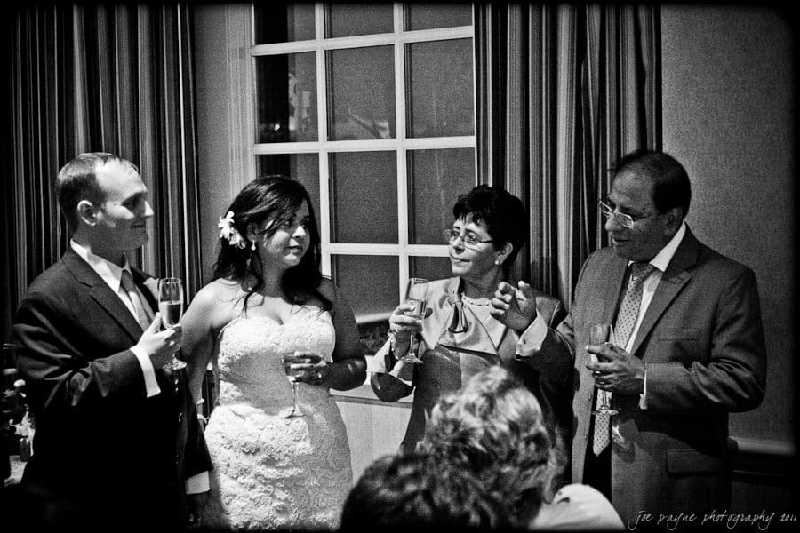 A black and white photo depicting four people standing indoors. A bride and groom hold champagne glasses next to two older individuals, also holding glasses, in conversation. The background shows a curtained window.