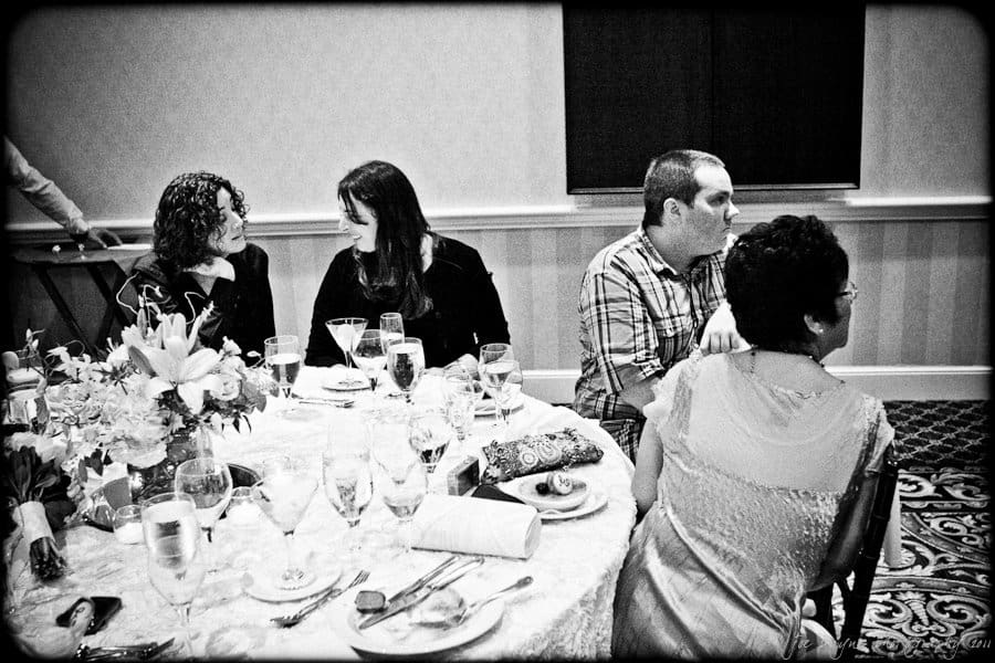 A black and white photo of four people sitting at a table during an event. The table is set with flowers, glasses, and plates. Two women are talking, while a man and another woman sit nearby, with patterned carpeting visible on the floor.