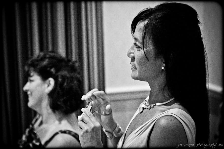 Black and white photo of two women indoors, smiling and standing side by side. The woman in the foreground holds a small object, possibly a phone. They are dressed elegantly, and a curtain is visible in the background.