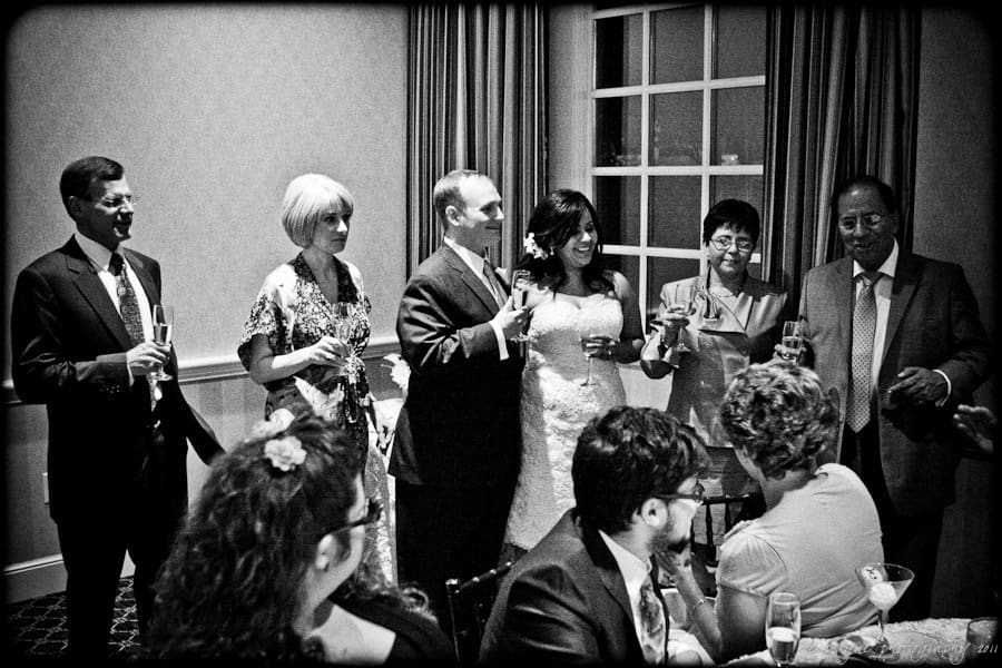 A black and white photo of a wedding reception where six people stand holding glasses, appearing to give a toast. A bride and groom are in the center. Seated guests in the foreground listen and watch the celebration.