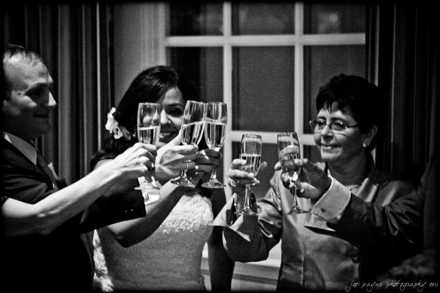 A group of people in formal attire raise champagne glasses in a toast. The setting appears to be indoors, with a window in the background. The image is in black and white, creating a classic and celebratory mood.
