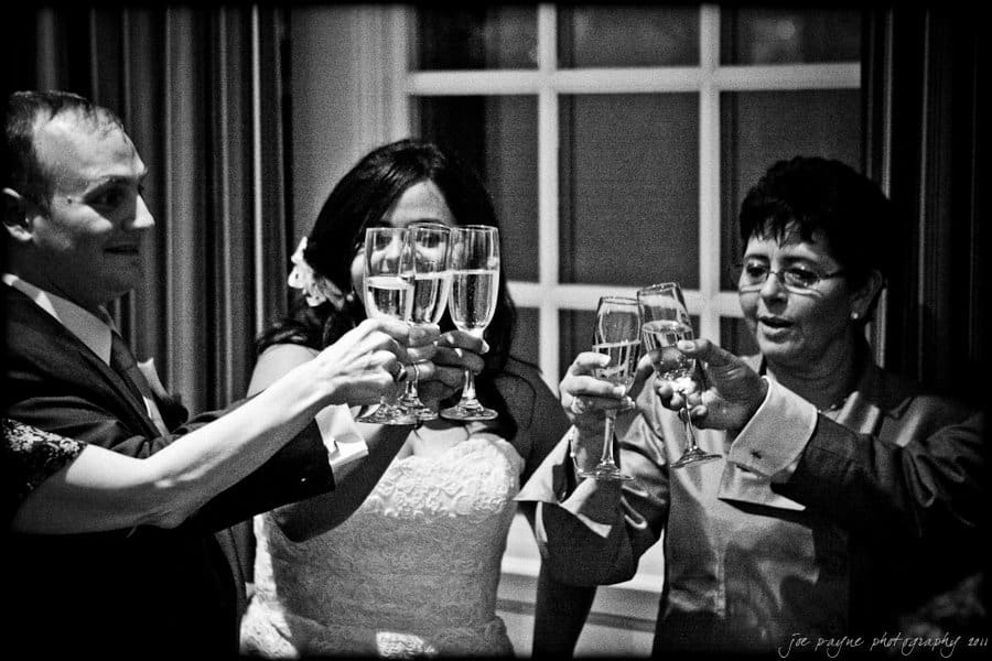 A group of people dressed in formal attire clinking champagne glasses together in celebration. The setting appears to be indoors, with a window in the background. The mood is joyous and festive.