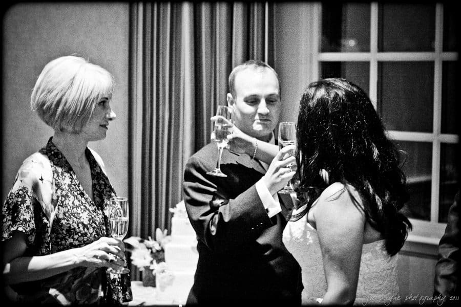 A black and white photo of three people standing together indoors, holding champagne glasses. The person in the center is in a tuxedo, interacting with a woman in a dress on the right. Another woman with short hair holds a glass on the left.