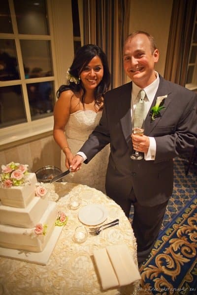 A bride and groom smile while cutting a three-tiered white wedding cake adorned with pink roses. The groom holds a glass of champagne. They stand in a warmly lit room near a window with curtains.