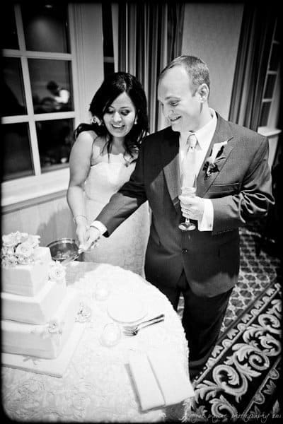 A bride and groom are smiling as they cut a tiered wedding cake together. The groom holds a champagne glass, and the bride looks at the cake. The scene is joyful, with a patterned carpet and draped curtains in the background.