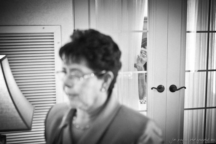 A black and white photo showing a woman out of focus in the foreground, wearing glasses and a jacket. In the background, another woman is smiling through a glass door, her face partially obscured by the curtain.