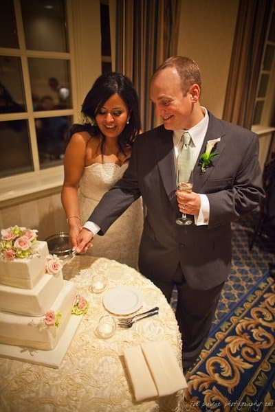 A bride and groom smile as they cut a three-tiered white wedding cake adorned with pink flowers. They stand beside a round table with lit candles in a warmly lit room with patterned carpet and curtains.
