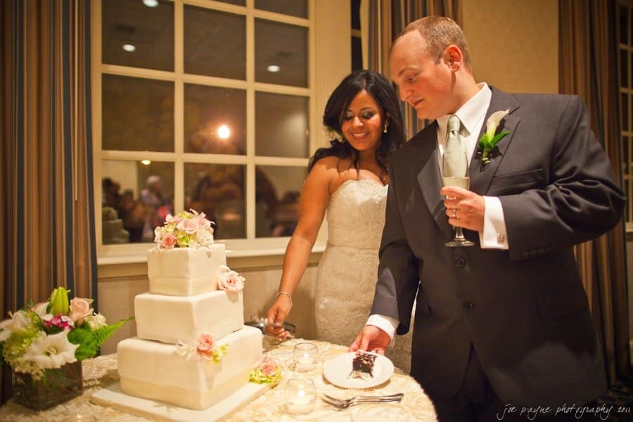 A bride and groom are standing next to a three-tiered white wedding cake with floral decorations. The groom cuts a piece while the bride smiles. They are in a warmly lit room with a large window in the background.