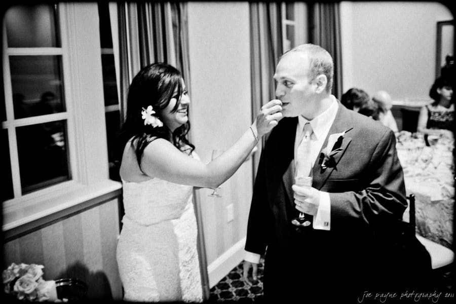 A bride feeds a piece of cake to the groom during a wedding reception. They are smiling and standing in a warmly lit room with curtains and a window in the background. Tables and guests can be seen in the background.