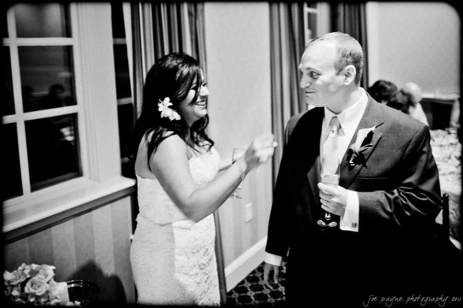 A bride and groom share a light-hearted moment indoors. The bride, wearing a strapless gown with a flower in her hair, laughs while the groom, in a suit and tie, holds a glass. They stand near a window with curtains.