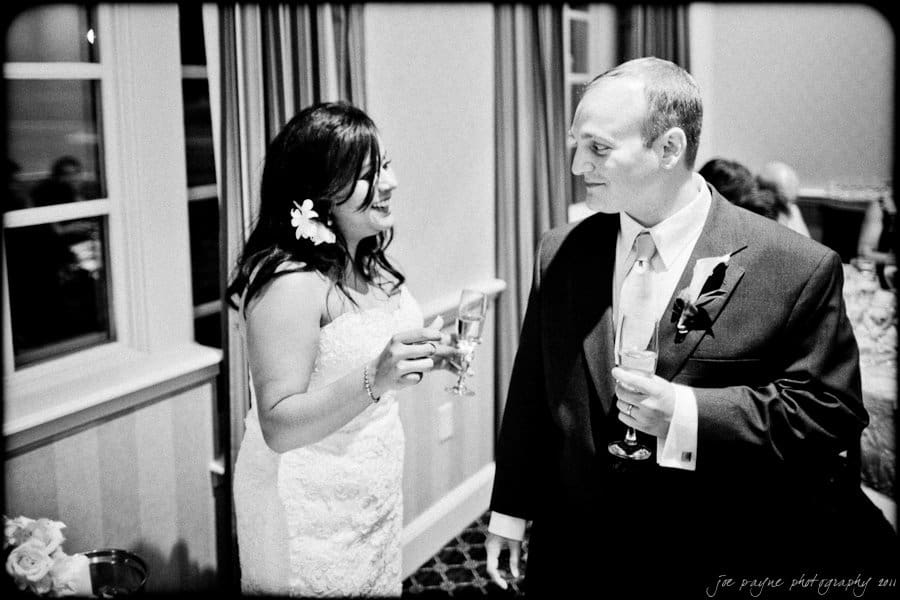 A bride and groom share a joyful moment, holding champagne glasses. She wears a white dress with flowers in her hair, and he is in a dark suit with a floral boutonniere. The setting appears to be indoors with soft lighting.