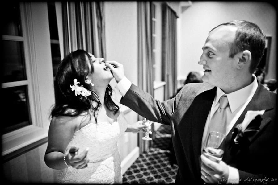 A couple in wedding attire playfully feeds each other cake. The bride laughs while the groom holds a small piece of cake. Theyre standing indoors, with a window and curtains in the background. The scene is captured in black and white.