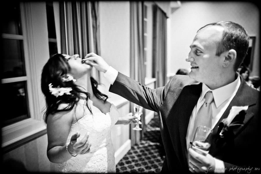 A bride and groom joyfully feed each other cake. The bride is wearing a white dress with flowers in her hair, and the groom is in a suit. They are smiling and holding champagne glasses, with a festive atmosphere around them.