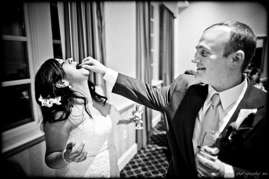 A groom playfully feeds the bride a piece of cake at their wedding. She is laughing while holding a glass, and he smiles, holding a glass in his other hand. The scene is joyful and celebratory, with decorations and curtains in the background.