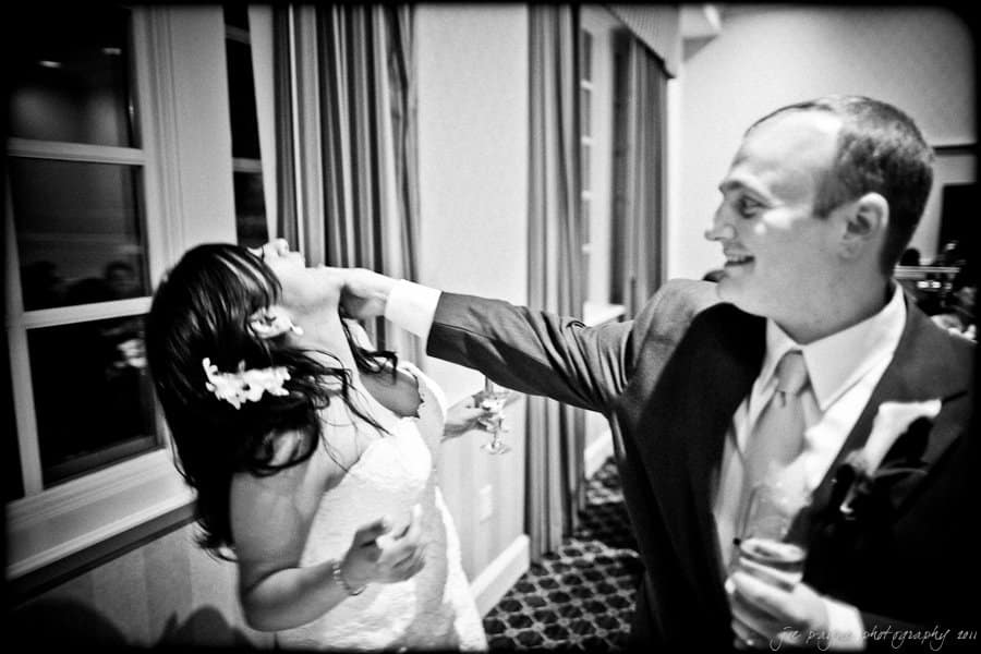 A bride and groom are playfully interacting at their wedding reception. The groom is gently touching the brides face, both smiling. They are dressed in formal attire, and the room is decorated elegantly.