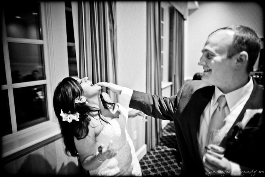 A bride and groom in a wedding setting, smiling and playfully interacting. The groom is gently feeding the bride a piece of cake. They are standing near a window with curtains, and the scene is joyous and lighthearted.