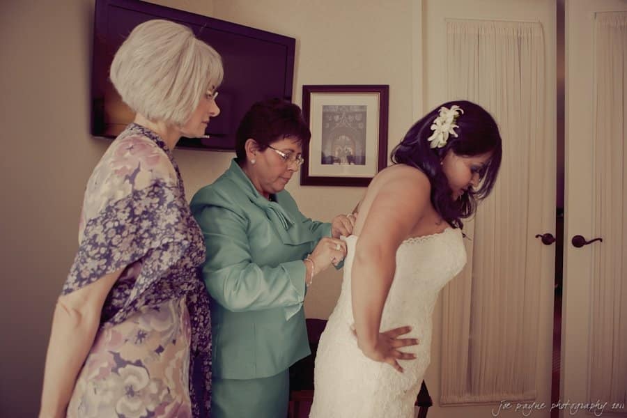 A bride in a white dress is being helped by two women, one in a blue suit and the other in a floral shawl. They are in a room with a TV and framed pictures on the wall. The bride has a flower in her hair.