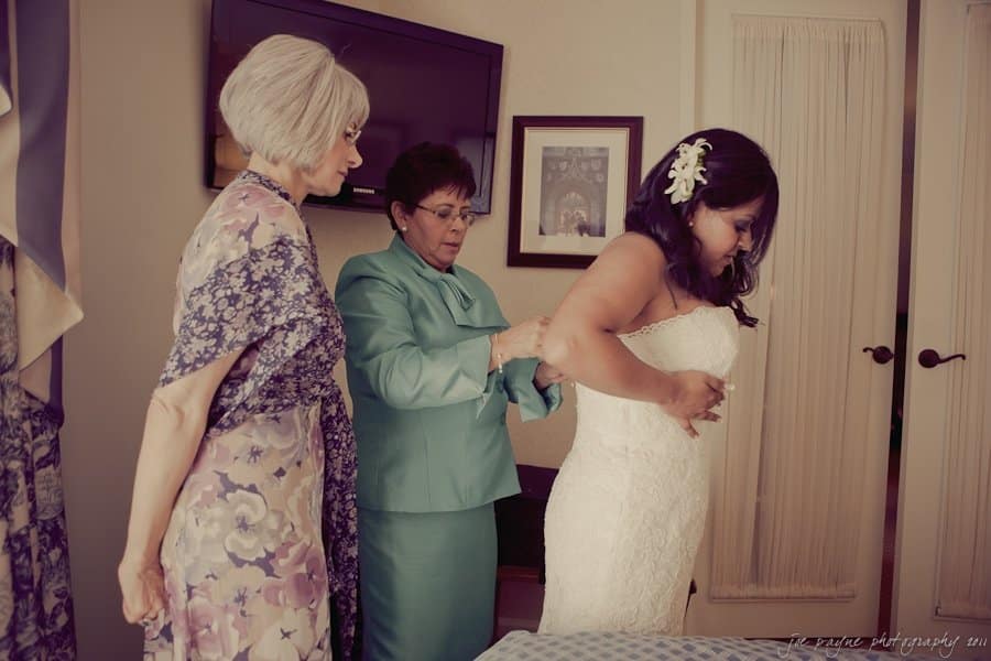 A bride in a white dress is being assisted by two women, one in a floral dress and the other in a teal outfit. They appear to be getting her ready for a wedding. The room has soft lighting and a framed picture on the wall.