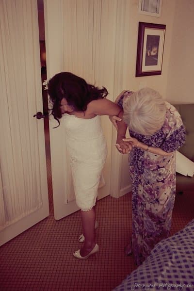 A bride in a white dress holds the arm of an older woman in a floral dress. They appear to be walking together in a warmly lit room with a bed in the foreground and a door in the background.