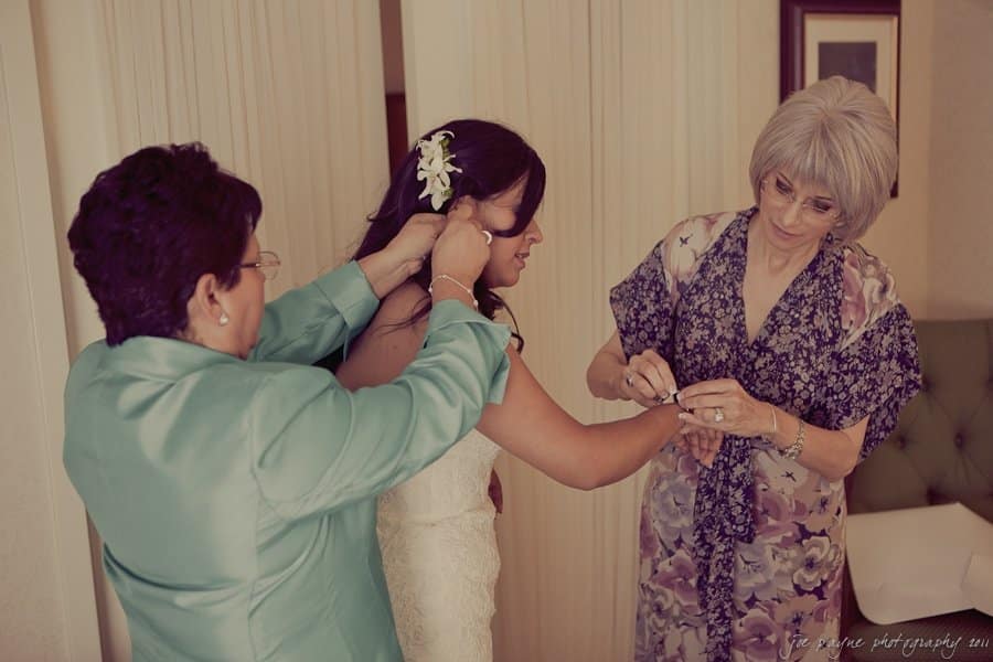 A bride in a white dress is assisted by two women, one adjusting her hair and the other fastening her bracelet. One woman wears a teal jacket, the other a floral dress. The scene is set in a softly lit room.