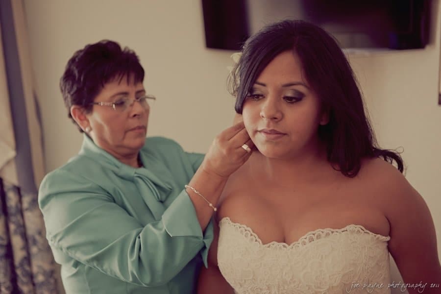 A bride in a strapless wedding dress receives assistance with her earrings from an older woman in a teal outfit. They are indoors, with a soft focus on the moment.