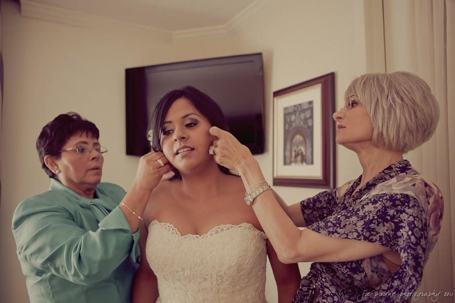 A bride in a strapless white gown is being assisted by two women. One woman adjusts her earring, and the other adjusts her veil. They are in a room with a wall-mounted TV and framed picture in the background.