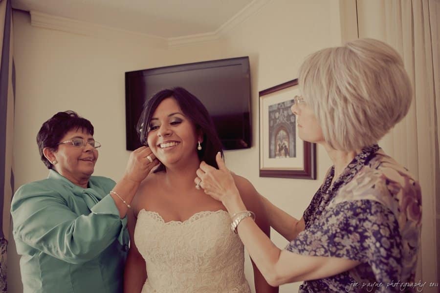 A bride smiles as two women, possibly her mother and a relative, help adjust her earrings and necklace. The bride is wearing a strapless lace wedding dress. They are in a room with framed photos on the wall.