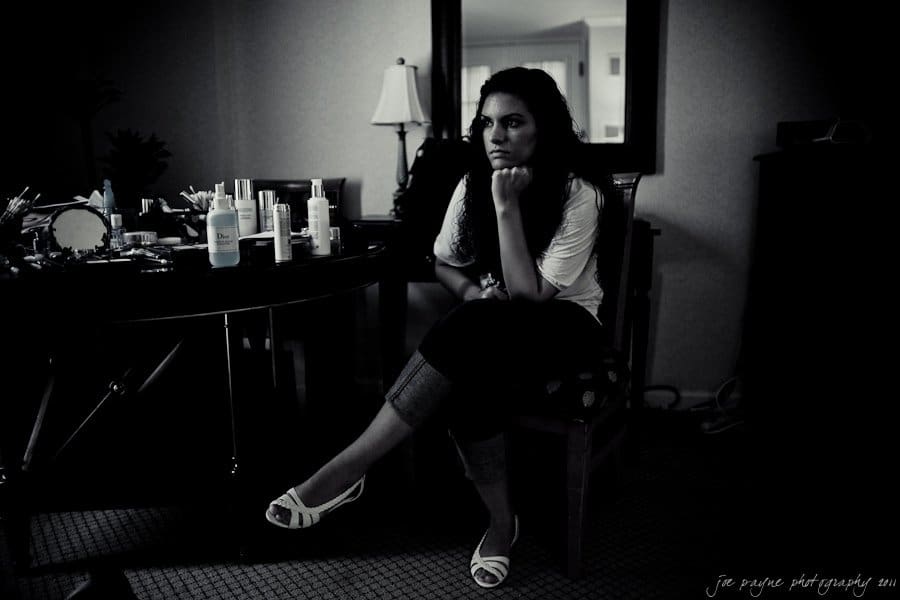 A woman sits in a dimly lit room, resting her chin on her hand. She is surrounded by various bottles and a mirror on a table. The mood is contemplative. A lamp, chair, and furniture are visible in the background.