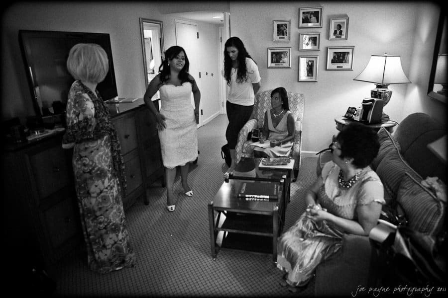 Black and white photo of a group of people in a living room, with a woman in a white dress standing, surrounded by others in formal attire. A lamp and framed pictures decorate the room, creating a cozy atmosphere.