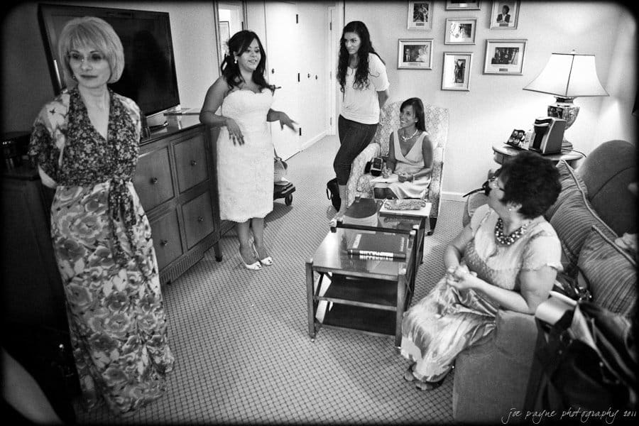 A black and white photo of a bride in a strapless dress standing in a living room with four other women, two seated and two standing. The room is cozy, with framed photos on the wall and a couch with a coffee table nearby.