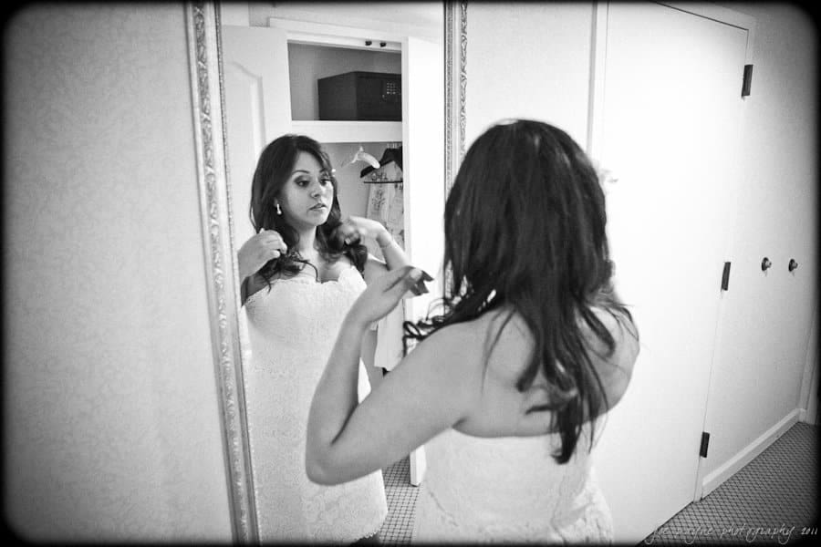 A woman stands in front of a mirror adjusting her earrings. She is wearing a strapless white dress and has long hair. The room has a closet in the background with an open door.