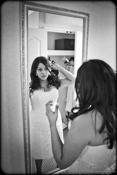 A woman in a white dress stands in front of a mirror, adjusting her hair. The reflection captures her focused expression. The room is softly lit, with an open closet visible in the background.