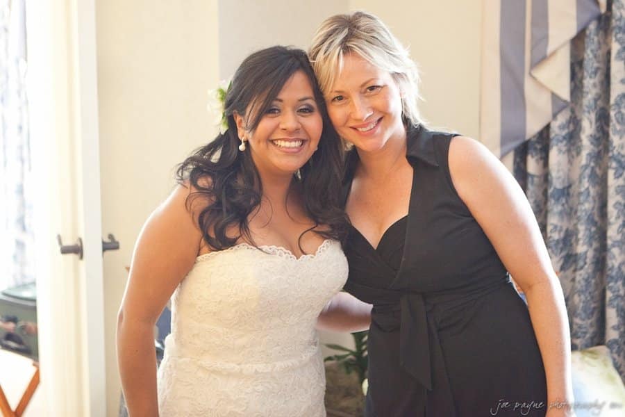 Two women are smiling and posing together. One is wearing a white lace wedding dress and the other a black sleeveless dress. They are standing in a well-lit room with floral curtains in the background.