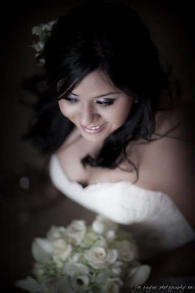 A bride with long dark hair and a strapless white gown smiles. She holds a bouquet of white flowers. The background is softly blurred, focusing on her face and upper body.