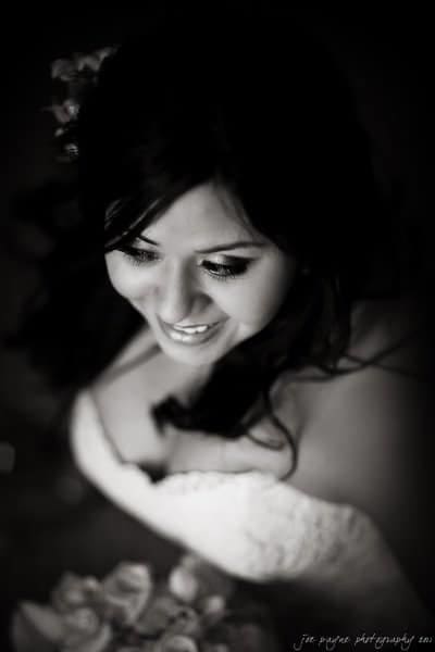 Black and white photo of a smiling bride with dark hair. The bride is looking slightly downwards, and her bouquet is visible at the bottom of the image. The photo focuses on her face and upper body.