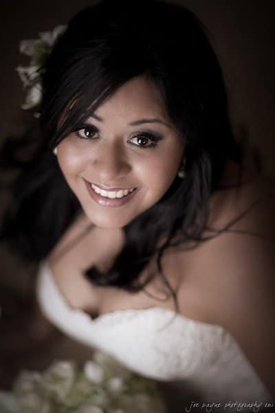 A bride with long dark hair smiles warmly at the camera, wearing a strapless white wedding dress. The soft-focus background emphasizes her joyful expression.