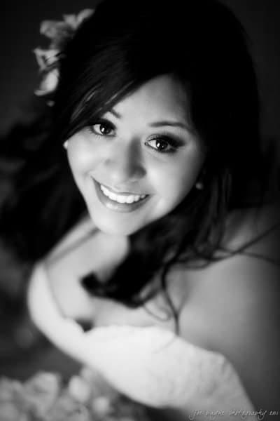 Black and white photo of a smiling bride looking up. She has long dark hair and wears a strapless wedding dress adorned with lace or floral designs. The focus is on her bright smile and expressive eyes.