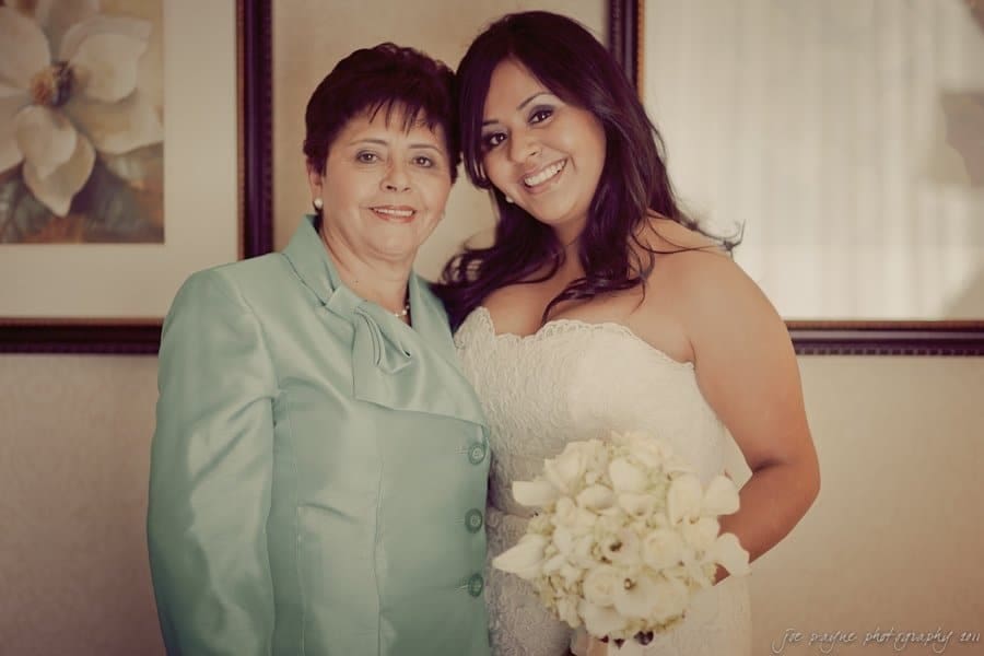 A bride in a white dress holds a bouquet of white flowers, standing beside a woman in a light blue outfit. They are smiling in a warmly lit room with floral artwork on the walls.