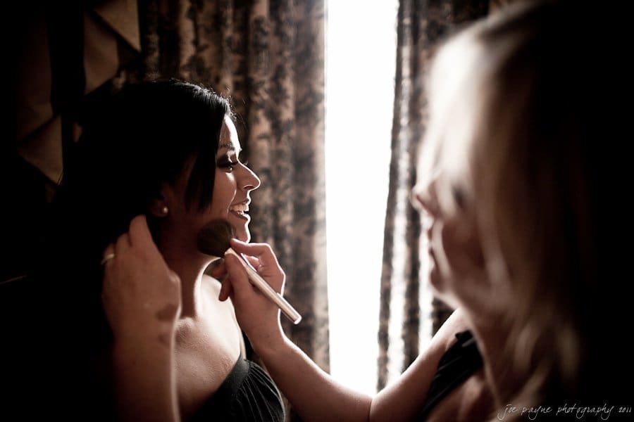 Two women are indoors, one applying makeup to the others face using a brush. They are surrounded by curtains and soft lighting, creating a warm atmosphere.