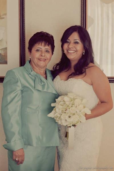 Two women are standing side by side, smiling. The woman on the right is in a white, strapless wedding dress holding a bouquet of white flowers. The woman on the left is wearing a light blue suit. They are in a warmly lit room with wall decor.
