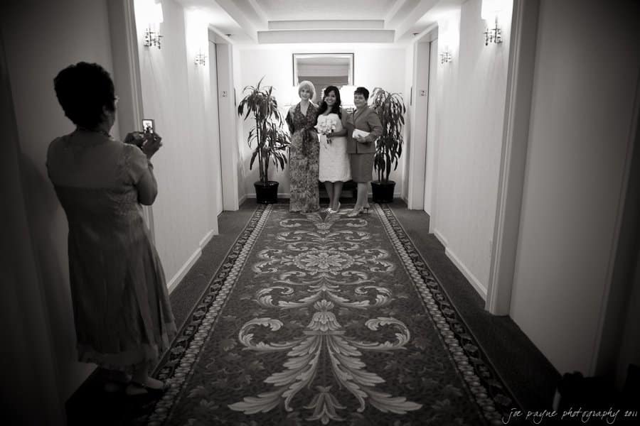 A black and white photo shows three women posing in a decorated hallway, with another woman taking a picture of them. They stand on an ornate carpet with plants beside them and lights on the wall.