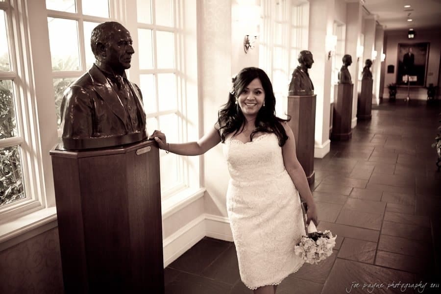 A woman in a white dress stands smiling in a hallway lined with busts on pedestals. She holds a bouquet in one hand and gently touches a bust with the other. The scene is warmly lit, and the floor tiles reflect the light.