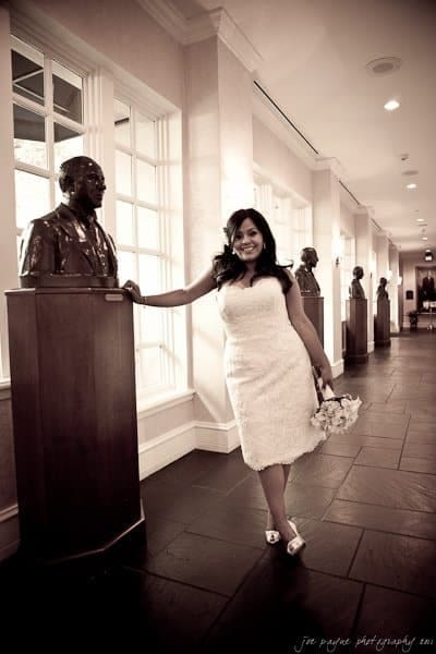 A woman in a white dress and holding a bouquet smiles while leaning against a bust in a hallway with other statues. The setting is elegant with tall windows and a tiled floor.