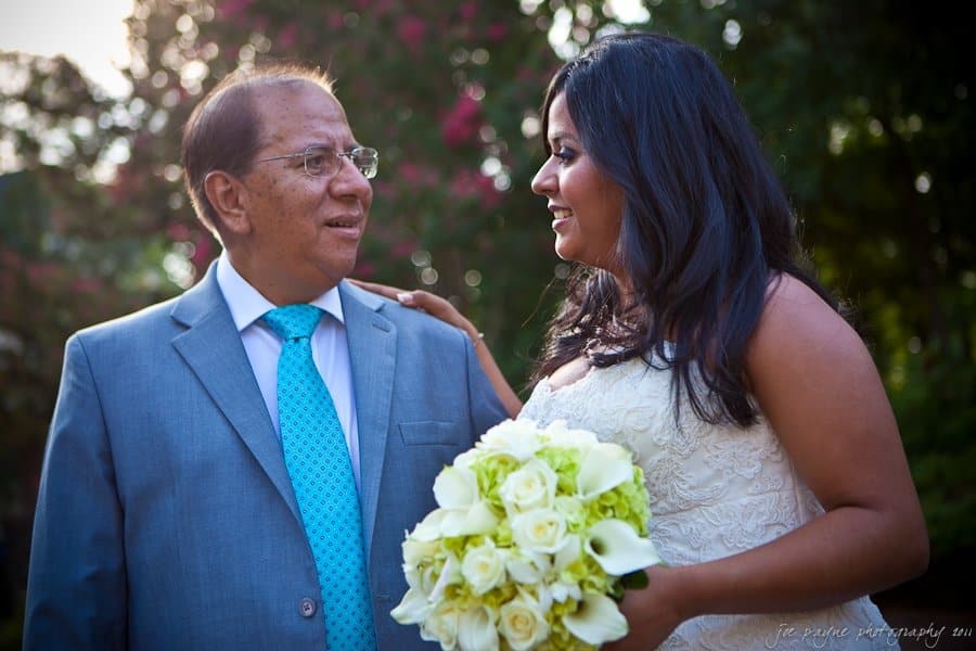 A bride in a white dress holds a bouquet of white flowers and gazes at an older man in a blue suit and tie. They are outdoors with greenery in the background. The atmosphere is warm and intimate.