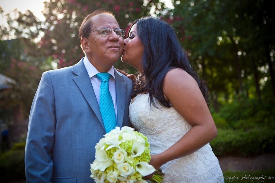 A bride in a white gown kisses an older man in a gray suit and blue tie on the cheek. She holds a bouquet of white roses. They stand outdoors with trees and sunlight in the background.