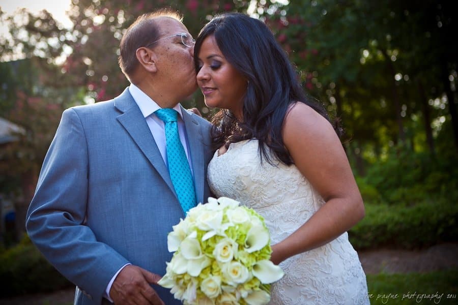 An elderly man in a gray suit kisses a woman in a white wedding dress on the cheek. She holds a bouquet of white flowers. They are outdoors, with greenery in the background.