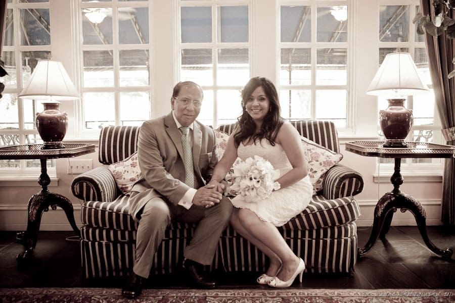 A man and woman sit on a striped sofa in an elegantly decorated room. The woman in a white dress holds a bouquet, while the man wears a suit. They are surrounded by lamps and windows, creating a warm atmosphere.