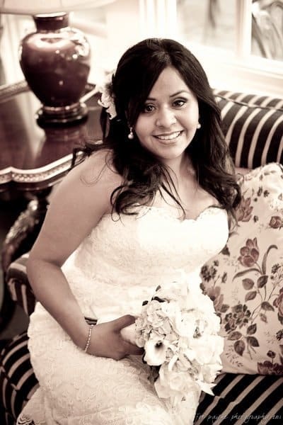 A smiling bride in a lace wedding dress holds a bouquet while sitting on a striped sofa with floral cushions. Background includes a decorative lamp on a table. Image in sepia tone.