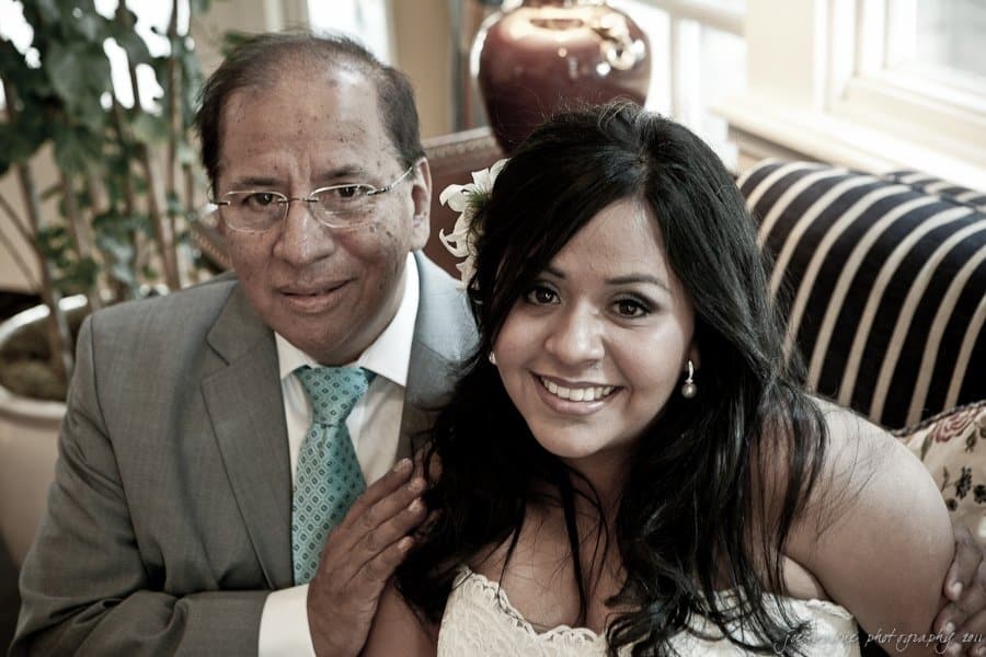 A smiling woman in a white dress with a flower in her hair sits beside an older man in a gray suit and teal tie. Theyre indoors, with a potted plant and a lamp in the background.